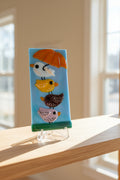 Fused glass panel with four stacked birds — white with orange umbrella, yellow, plum-brown, and pink — on sky blue background with green grass base, displayed on clear acrylic stand