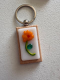 Orange flower fused glass keychain with red center, green stem, and teal leaf on white glass tile with wood backing and silver key ring