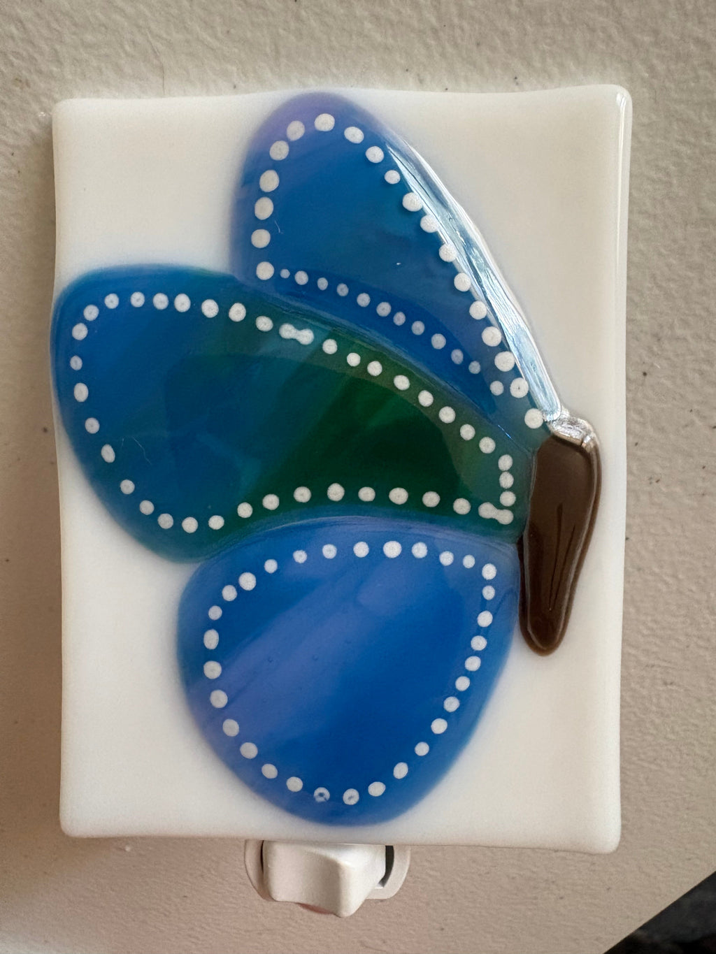 Blue and green fused glass butterfly nightlight glowing warm when plugged in, showing cobalt and emerald wings with white dot detailing