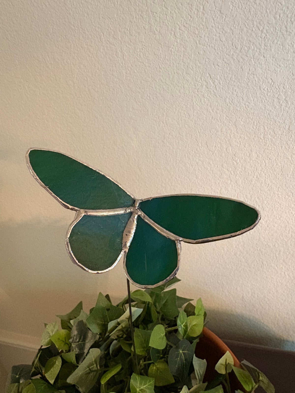 Emerald green and teal iridescent stained glass butterfly plant stake displayed in a leafy planter